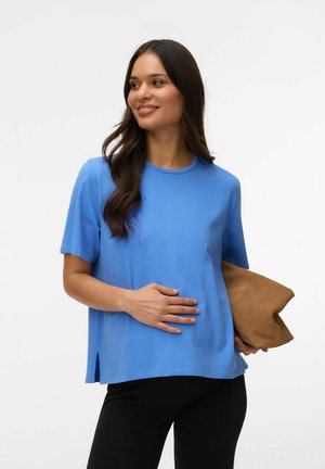 Kvinde med langt mørkt hår iført en blå kortærmet skjorte og sorte bukser, der holder en brun clutch og smiler, mens hun kigger sidelæns.