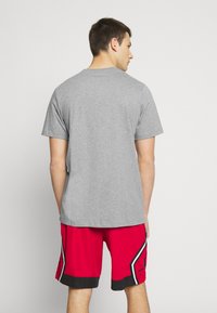 Camiseta de algodón gris con mangas cortas, combinada con pantalones cortos deportivos rojos que tienen acentos en negro y blanco y un logo en la pierna izquierda.