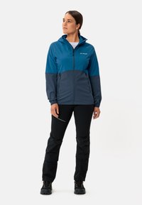 Veste zippée bleu et gris avec capuche ; conçue en matériau léger et résistant à l'eau. Pantalons noirs avec panneaux texturés.
