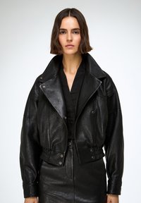 Mujer con cabello corto y castaño, vestida con una chaqueta de cuero negra y falda a juego, de pie contra un fondo claro y sencillo.