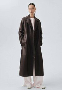 Brązowy skórzany długi płaszcz z wyciętym kołnierzem, bocznymi kieszeniami i otwartym przodem, noszony na jasnoróżowym stroju, z gładko uczesanymi włosami i wyrazistymi kolczykami.