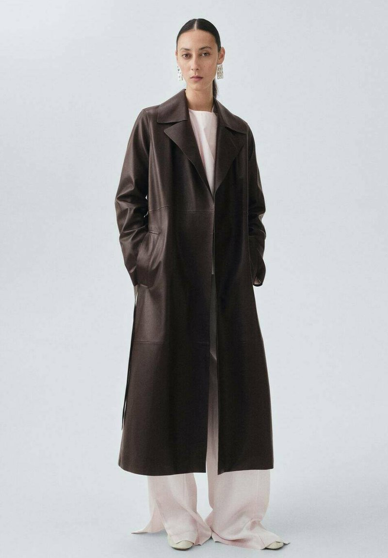 Brązowy skórzany długi płaszcz z wyciętym kołnierzem, bocznymi kieszeniami i otwartym przodem, noszony na jasnoróżowym stroju, z gładko uczesanymi włosami i wyrazistymi kolczykami.