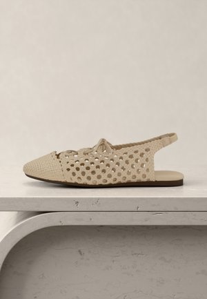 Beige woven flat sandal with open sides and back strap, featuring a lace tie on top, placed on a light grey marble surface.