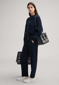 Marineblauer Trainingsanzug mit einer Jacke zum Zipper, passenden Hosen und weißen Sneakern. Enthält eine gemusterte Handtasche in Schwarz und Gold als Accessoire.
