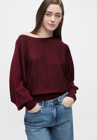 Bordowy, oversize sweter z dzianiny z szerokim dekoltem, długimi rękawami i ściągaczami, w zestawieniu z jasnoniebieskimi, wysokim stanem jeansami.