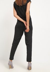 Svart jumpsuit med korta ärmar, skräddarsydd passform och tapered ben, matchad med ljusgrå klackade ankelskor med dragkedjedetalj.