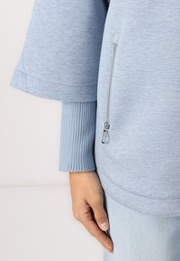 Lyseblå oversized sweatshirt med teksturert stoff, en glidelåslomme og ribbede mansjetter for ekstra detaljer og kontrast.