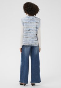 Egy göndör, sötét hajú nő hátulról látható, aki ujjatlan, kékesszürke kötött mellényt visel egy fehér, hosszú ujjú ing fölött, valamint bő lábú kék farmernadrágot.