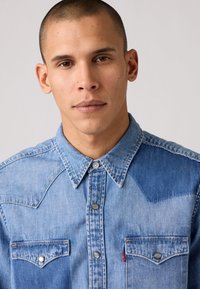 Camisa de mezclilla azul clara con botón en la parte delantera, dos bolsillos en el pecho, un cuello con punta y paneles en contraste en los hombros.