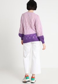 Stickad tröja i ljuslila med diamantformade utskärningar, med en mörkare lila fåll. I kombination med vita vida byxor och färgglada sneakers.