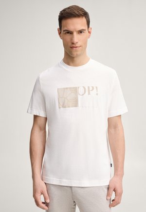 Homme debout, portant un t-shirt blanc avec un rectangle beige à motifs et le texte « OP! », fond neutre, mains le long du corps, face à l'avant.