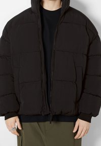 Svart puffjacka med dragkedja, tjocka quiltade paneler och huva. Har elastiska manschetter och en avslappnad passform. Bärs över en svart skjorta.