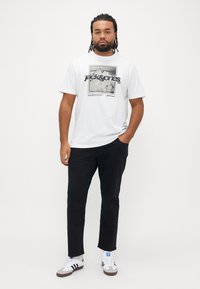Λευκό βαμβακερό t-shirt με γραφικό σχέδιο που απεικονίζει βουνά και το κείμενο "jack&jones". Συνδυασμένο με μαύρο παντελόνι και λευκά αθλητικά παπούτσια.