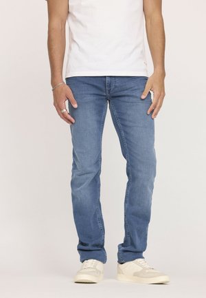 Jean en denim bleu clair avec une coupe droite, détail de whiskering léger et zip. Associés avec des baskets blanches.