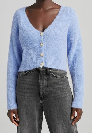 Persoon draagt een lichtblauwe, zacht uitziende, knoopjesvest met lange mouwen en zwartvervaagde jeans met hoge taille.