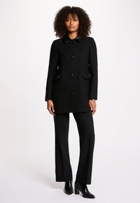 Schwarzer Mantel mit Kragen, Knöpfen auf der Vorderseite und seitlichen Taschen; kombiniert mit schwarzen Hosen und Stiefeln mit Absatz. Glatte Textur mit taillierter Passform.