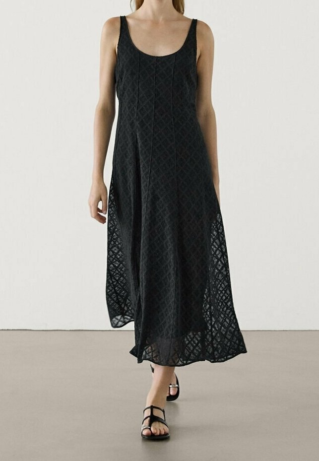 Femme portant une robe maxi noire sans manches avec un motif diamant texturé et des sandales plates noires à brides, marchant sur un sol lisse.