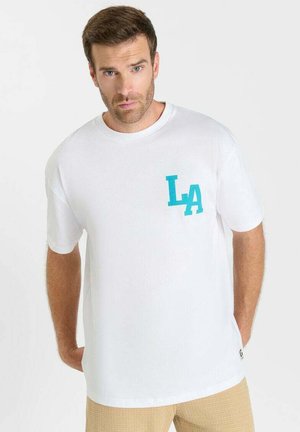 Maglietta bianca con lettere turchesi "LA" sul petto indossata da uomo in piedi con capelli castani corti e barba, mani dietro la schiena.