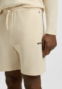 Vaaleanbeige teksturoidut shortsit, joissa on kiristysvyötärö ja musta "BOSS"-logo sivussa. Kangas on vaahtomuovimainen.