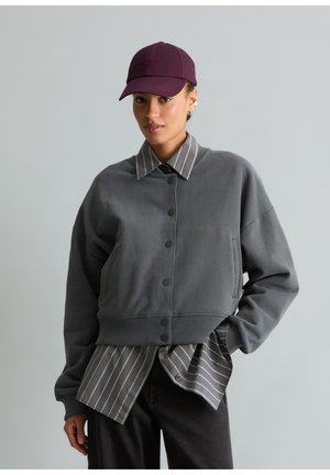 Person trägt eine dunkelgraue, geknöpfte Jacke über einem grauen, fein gestreiften Hemd, schwarze Hose und eine bordeauxrote Baseballkappe, steht vor einem einfarbigen Hintergrund.