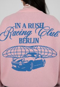 Pull en tricot rose avec un texte graphique bleu "IN A RUSH Racing Club BERLIN" et une illustration d'une voiture avec un globe au-dessus.