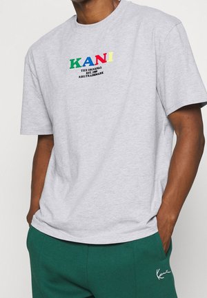 Mężczyzna ubrany w jasnoszary t-shirt z kolorowym napisem „KANI” i czarnym sloganem, w połączeniu z zielonymi spodniami z białym haftowanym logo.