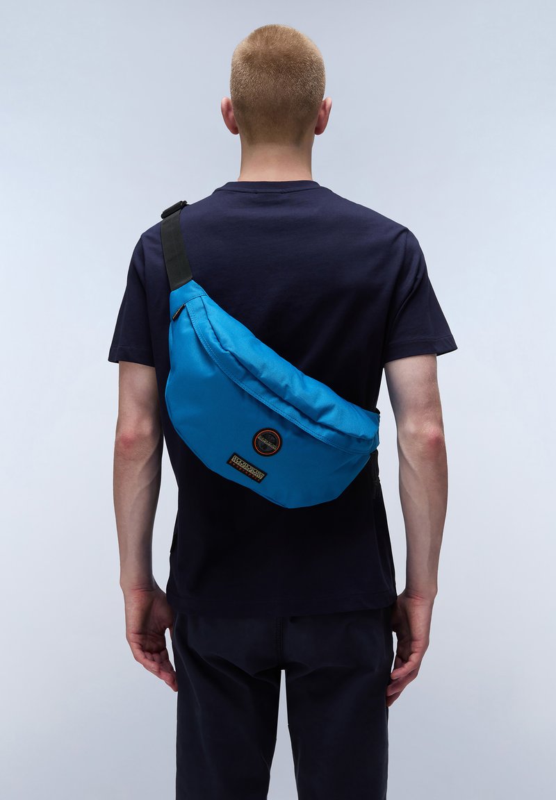 Blå nylonaxelväska som bärs över kroppen, med en böjd design, en logopatch och ett justerbart svart band.