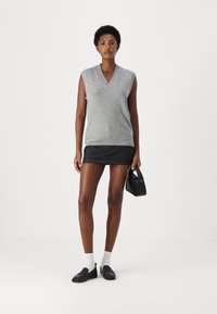 Pull gris sans manches avec un décolleté en V, associé à une mini-jupe noire. Le mannequin tient un petit sac à main noir, portant des chaussettes blanches et des mocassins noirs.