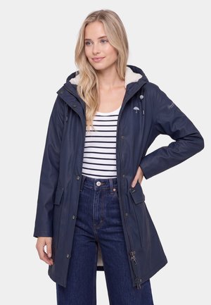 Vrouw met lang blond haar die een marineblauwe kapmantel draagt over een gestreept shirt en blauwe spijkerbroek, staand met één hand op haar heup tegen een witte achtergrond.