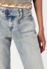 Jeans de mezclilla de lavado claro con cintura alta, que cuentan con dos botones de metal, un bolsillo frontal y un ligero desgastado a lo largo del muslo y la parte trasera.
