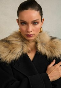 Melns vilnas mētelis ar gaiši brūnu kažokādu apkakli, ar dubultu pogu slēgumu un teksturētu apdari, uzsverot greznos akcentus.