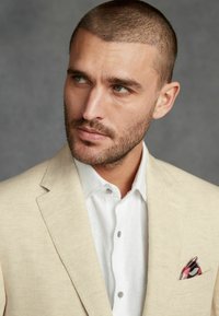 Blazer en lin beige sur une chemise blanche, avec un pochette en soie multicolore. Gros plan mettant en avant la texture du tissu et la coupe ajustée.