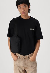 Zwarte schoudertas met ritszakken, gedragen over een zwart T-shirt en beige broek met een gestructureerde afwerking. Eenvoudig ontwerp en praktische stijl.