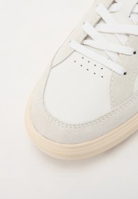 White leather and grey suede sneaker with a rounded toe, contrasting stitching, and white laces, featuring small ventilation holes.