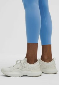 White athletic shoes with mesh detailing, round toes, and a textured sole, paired with fitted light blue leggings, showing a close-up perspective.