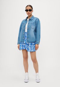 Jeans jakna svetlo modre barve, z gumbom in sprednjim žepom, v kombinaciji z nabranim krilom z modrim in belim vzorcem ter belimi supergami.