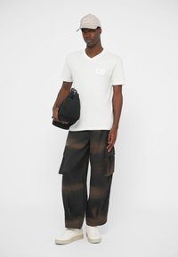 Biela bavlnená tričko s výstrihom do V a malým logom, v kombinácii s čiernymi cargo nohavicami s hnedým pruhovaným vzorom. Obsahuje čiernu tašku a béžovú čiapku.