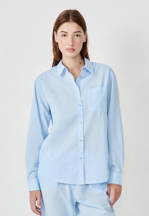 Giovane donna con lunghi capelli castani che indossa una camicia azzurra chiara con bottoni e pantaloni coordinati, davanti a uno sfondo semplice e chiaro.