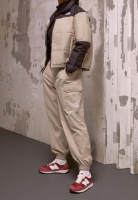 Flera jacka i beige och svart, beige cargobyxor, röda och vita sneakers. Slät tyg, elastiska ärmar och synliga logodetaljer.