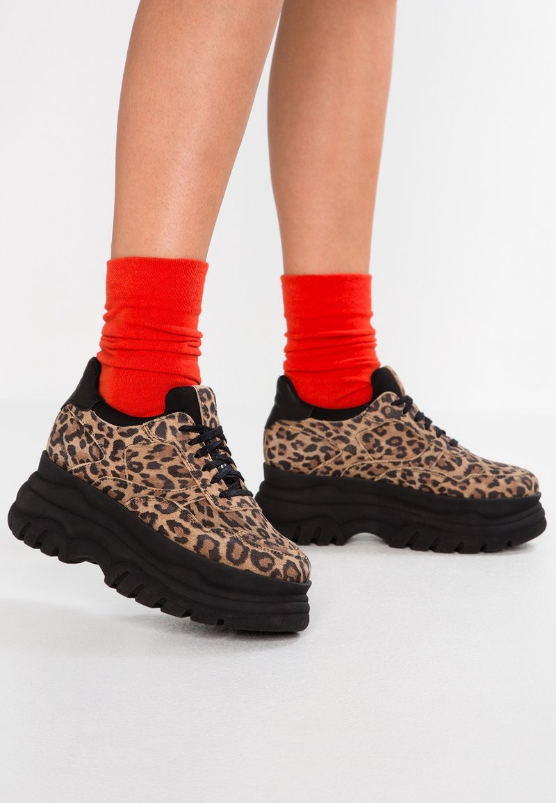 Baskets imprimées léopard avec une semelle noire épaisse, dotées d'un design à lacets et de chaussettes rouges contrastantes. Texture semblable à du daim avec des motifs texturés.