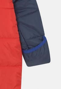 Czerwona rękaw kurtki puchowej z granatową tkaniną, z marszczonym mankietem, niebieską ozdobną taśmą i pikowanym wzorem zapewniającym izolację.