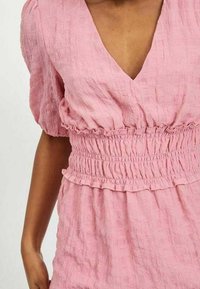 Rosa texturerad klänning med V-ringning och korta puffärmar; har en rynkad midja och volangdetalj i nederkant, tillverkad av lättviktsmaterial.