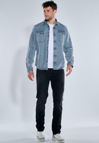 Hellblaues Jeanshemd mit zwei Brusttaschen, Knopfleiste, über einem weißen T-Shirt getragen, kombiniert mit schwarzen Hosen und beigen Sneakern.