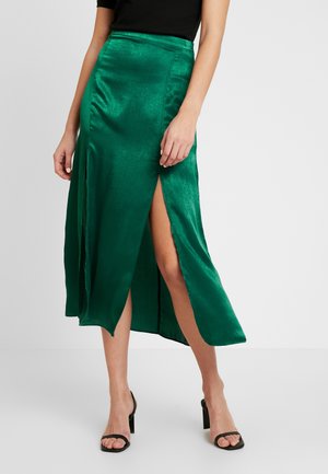 Mujer con una falda midi verde brillante con aberturas laterales combinada con sandalias negras de tacón alto y una camiseta negra, de pie sobre un fondo liso.