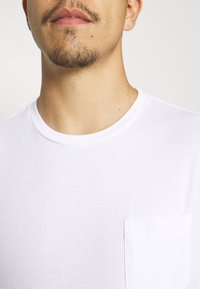 T-shirt bianco in cotone con scollo rotondo, dotato di una piccola tasca a sinistra sul petto e una texture liscia e leggera.