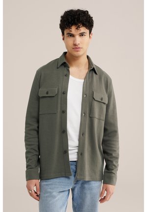 Tânăr cu păr creț, purtând o cămașă verde măslinie cu nasturi peste un tricou alb și blugi bleu deschis, stând în picioare pe un fundal simplu.