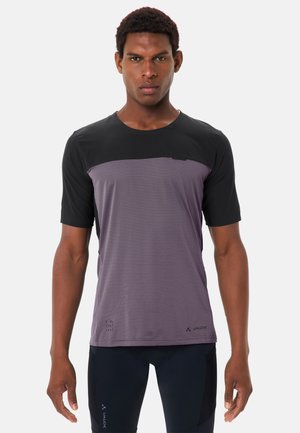 Mann trägt ein schwarzes und violettes kurzärmeliges Radtrikot und schwarze Radhosen, steht vor einem schlichten weißen Hintergrund.