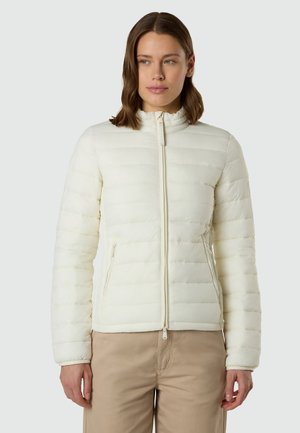 Donna con capelli castani alla lunghezza delle spalle che indossa una giacca trapuntata bianca con zip e pantaloni beige, in piedi davanti a uno sfondo grigio chiaro.