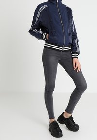 Námořnická bomber bunda s bílými bočními pruhy, předním zipem a žebrovanými manžetami. V kombinaci s tmavě šedými skinny džínami a černými masivními teniskami.