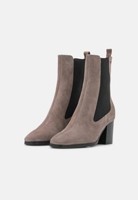 Bottines en suede taupe avec des panneaux latéraux en élastique noir et des talons bloc empilés, présentées sur un fond blanc.